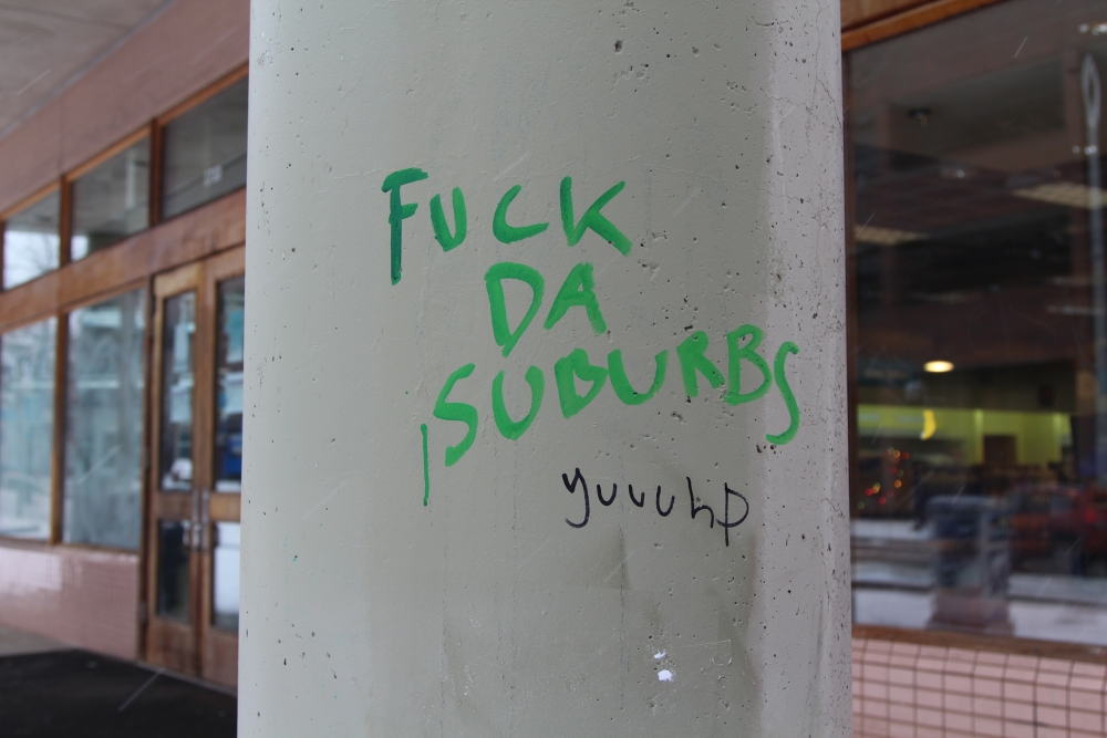 Gray cement pillar with "Fuck Da Suburbs" written on it in green