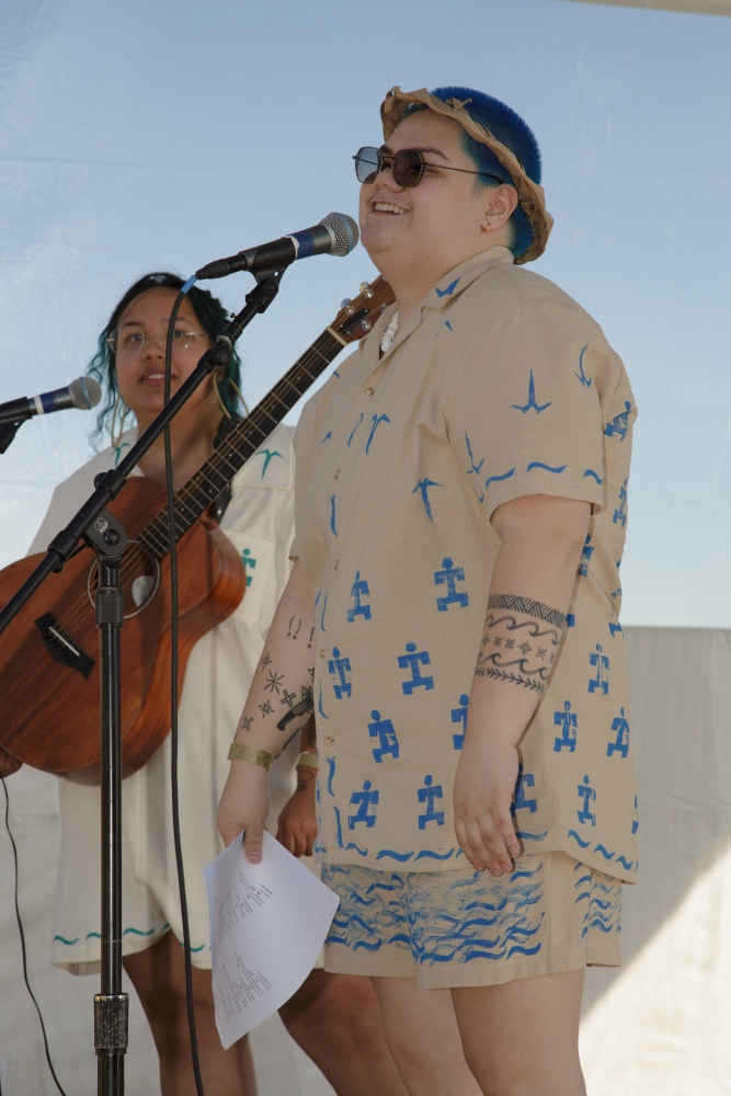 Young woman with long dark hair and glasses plays guitar next to a singer dressed in blue cap, sunglasses, and beige shirt with indigenous-style icons print.