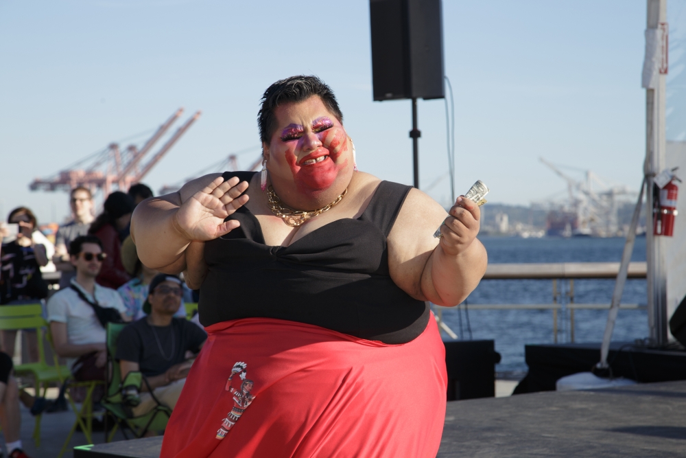 Indigenous woman with short black hair and red face paint, in black bodice top and red skirt, dances and smiles against sunny waterside backdrop.