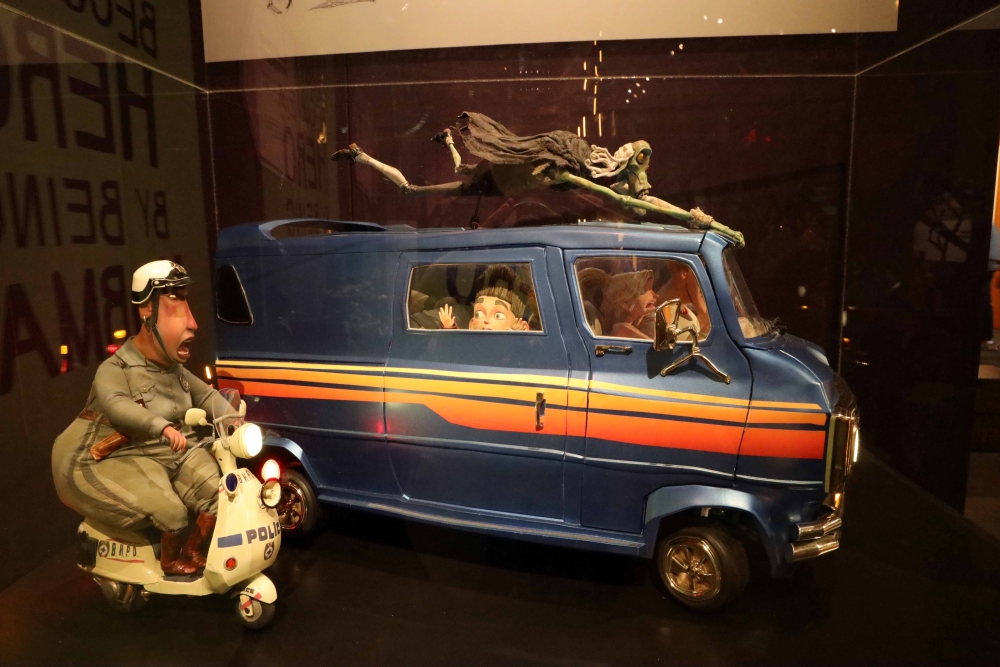 Sculpture of minivan with woman driver and worried-looking child's face at a passenger window; a thin, green monster figure hovers above the van, which is flanked by a hefty-looking police officer on a motorbike.