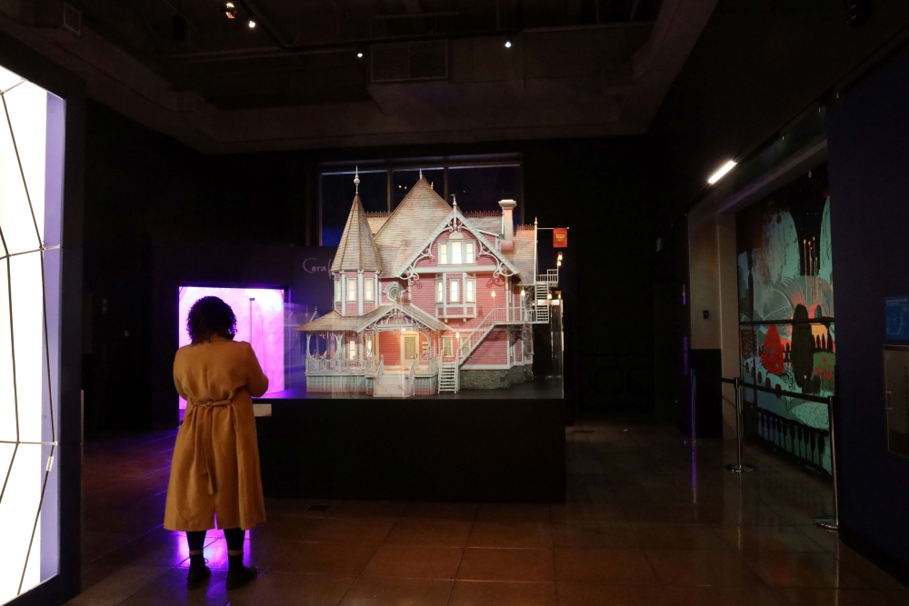 Woman in light-brown coat, seen from behind, stands in front of lit-up mockup of fanciful pink house with turrets and gables.