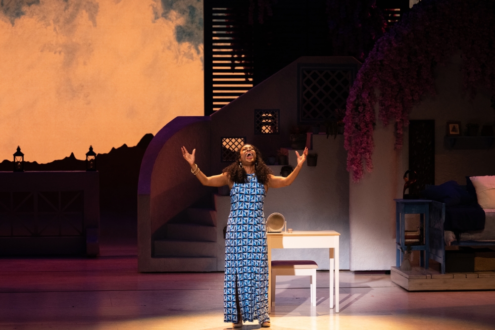 Lisa Estridge, a tall Black woman with long, pulled-back curly hair belts out a solo number in the middle of the stage while wearing a blue and white patterned dress.