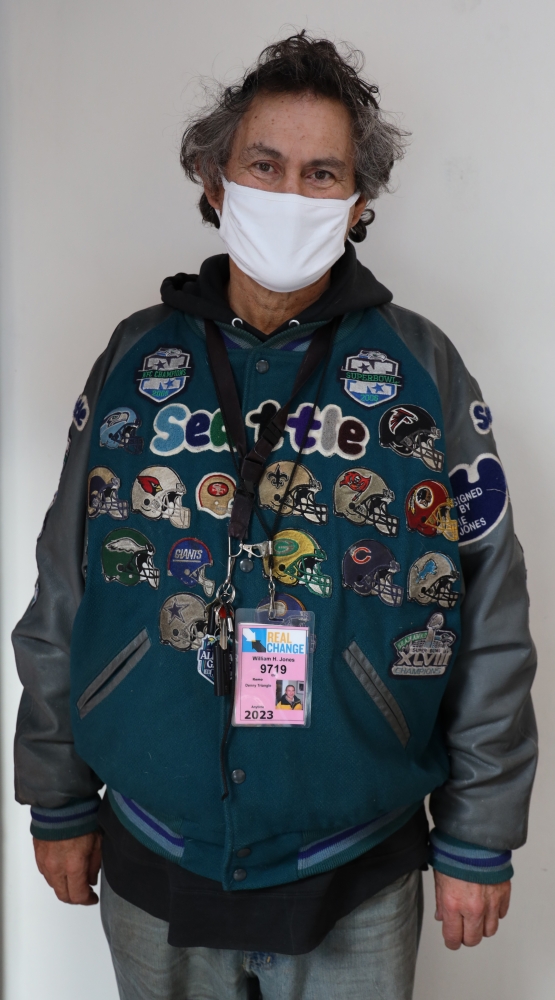 Middle-aged white man wearing white face mask and blue jacket covered with Seattle sports logos