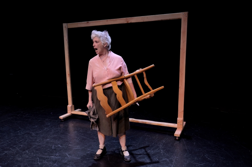 Older white woman with short hair, dressed in pink blouse, gray skirt, bobby socks, and Mary Janes, stands in front of a wooden frame, holding a chair, mouth open.