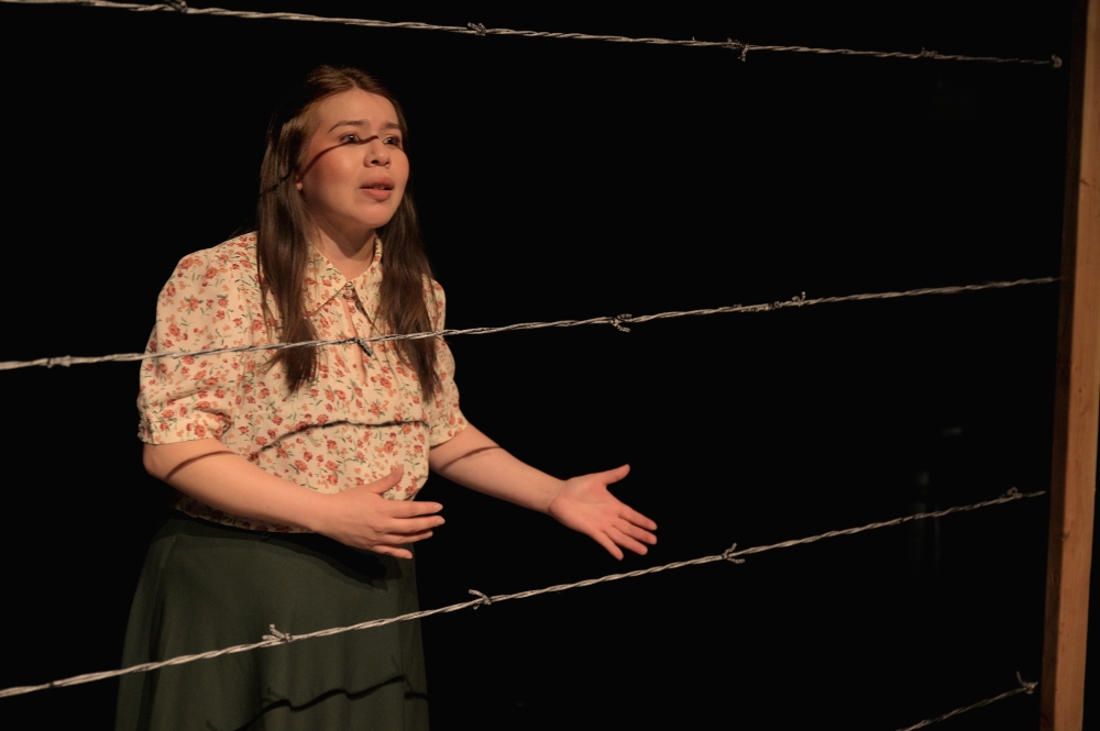 Young Latina-looking woman in flower-print blouse and green skirt stands behind line of barbed wire.