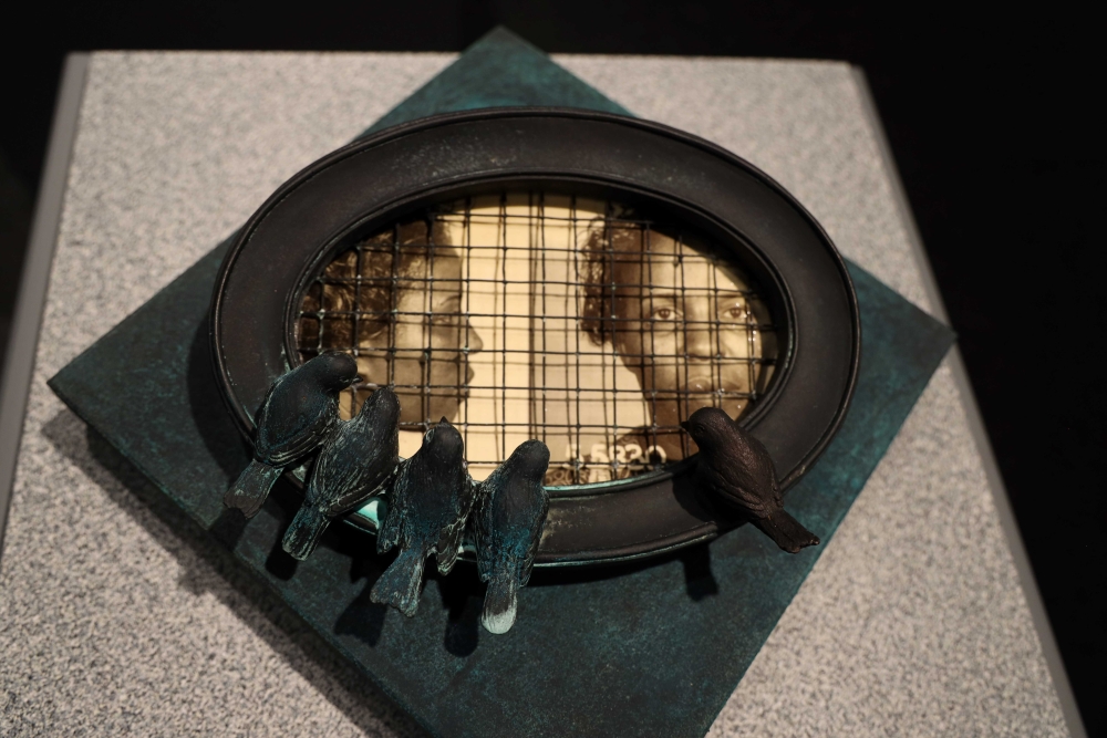 Sculpture showing five birds perched on a metal rim enclosing a grid behind which is visible two photographs of a Black woman with curly hair, one taken from the side and one from the front