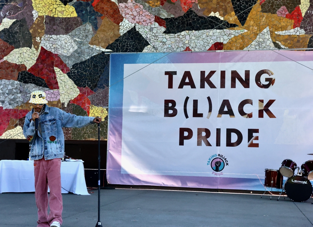 Man in cap and jeans stands at mic in front of colorful abstract mural and sign that says, "Taking B(l)ack Pride."
