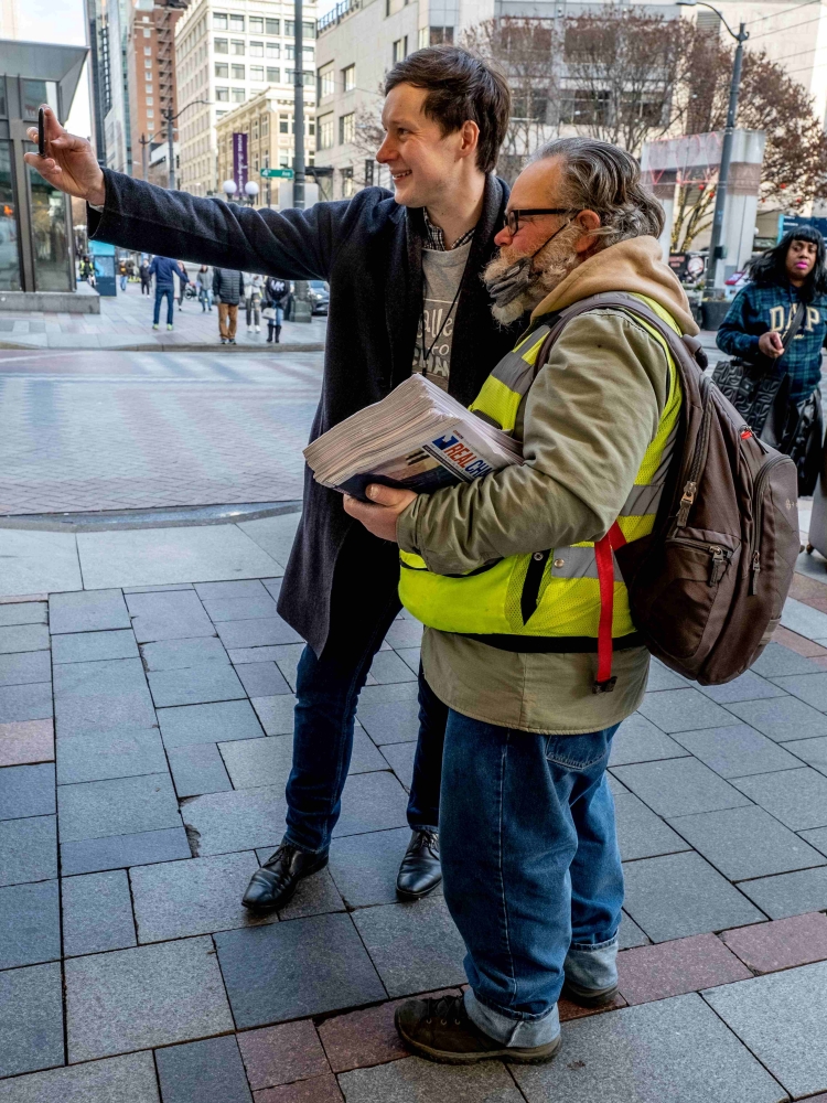 Man in dark jacket and pants holds up phone to take picture of himself and older man in Real Change vendor yellow jacket.
