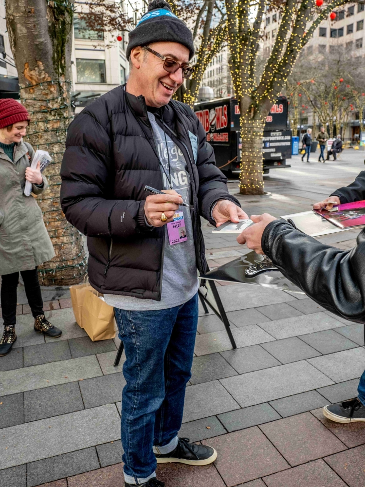 Middle-aged white man in glasses, puffy jacket, and knit hat smiles while signing a CD.
