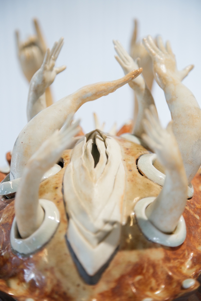 Ceramic vulva set in an orange mound, out of which a circle of arms and hands reach out around it.