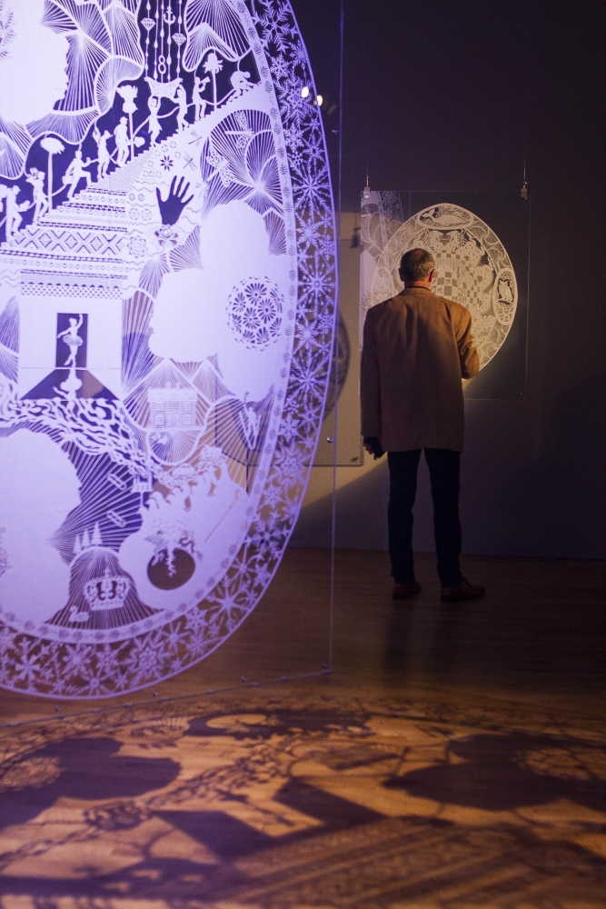 Foreground shows ornate paper cut-out, colored purple, featuring flowers, silhouetted figures, and handprints