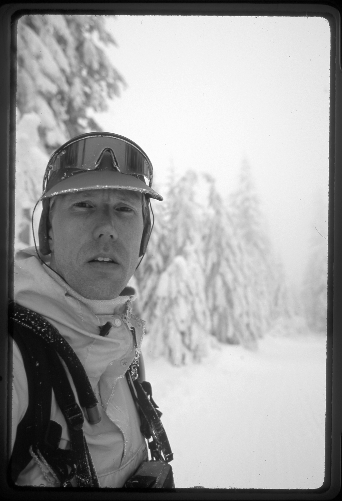 Young white man with snow goggles on head, seen against snowy backdrop with skis